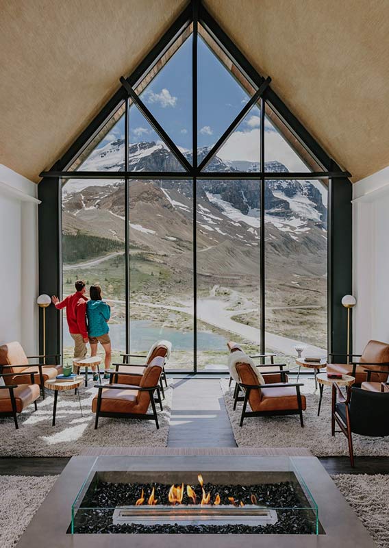Two people stand at a large window overlooking mountains.